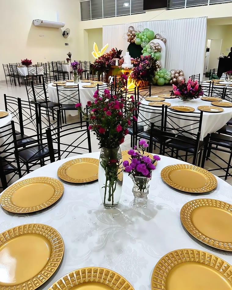 Salão montado para evento formal, com toalhas brancas, pratos dourados e arranjos de flores