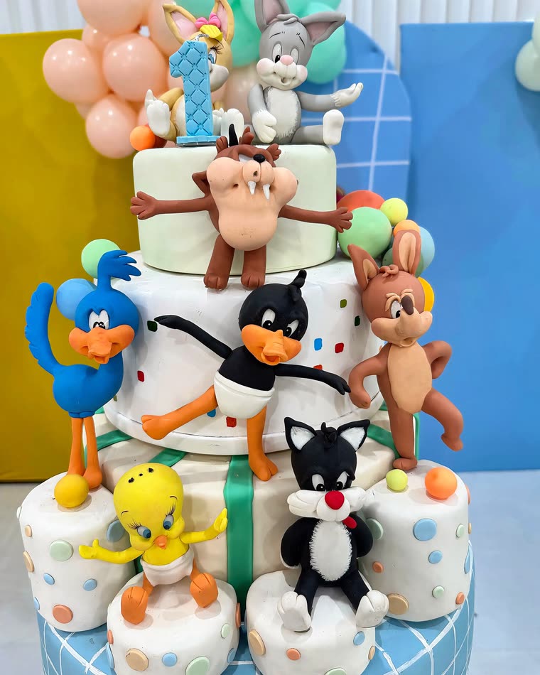 Bolo de aniversário infantil de vários andares, com os personagens da festa e painéis de balões ao fundo