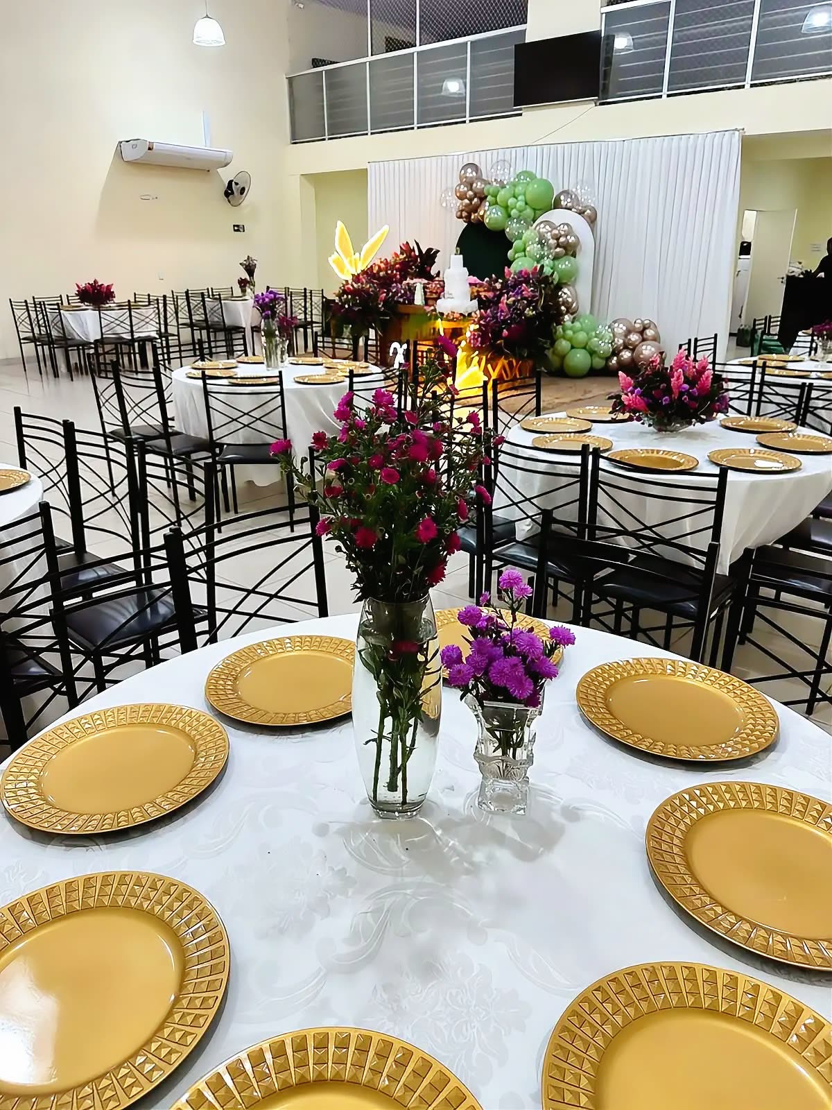 Salão montado para evento formal, com toalhas brancas, sousplat dourado e arranjos.